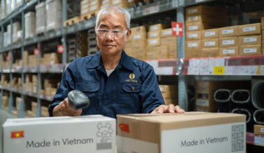 AI generated image of an Asian man in a warehouse with several shelves of cardboard boxes behind him as he scans two boxes in front of him getting ready to ship. Boxes say made in Vietnam.