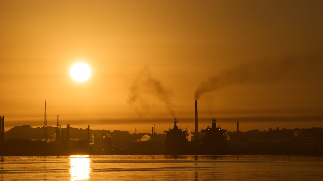 An sun hangs low in an orange sky over smoke stacks, with plumes of smoke rising from them, at an oil refinery in Havana Bay, Cuba