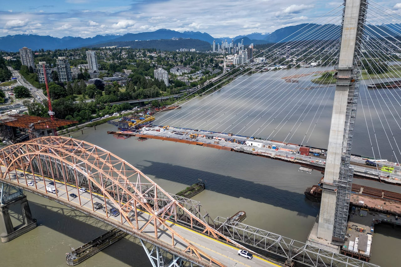 An aerial shot of an orange bridge with cars on it is to the left of an under-construction bridge over a river.