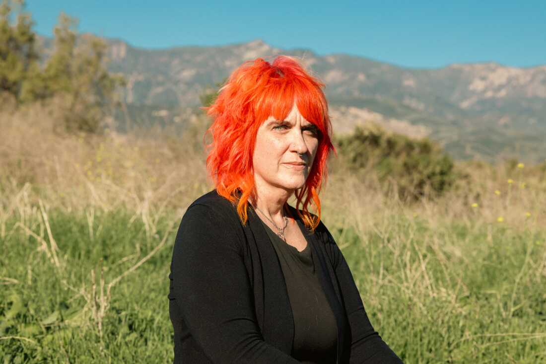 Micky Small, 53, is a screenwriter and is one of hundreds of millions of people who regularly use AI chatbots. She has bright orange, shoulder length hair and is seated with a background of mountains and brush behind her.