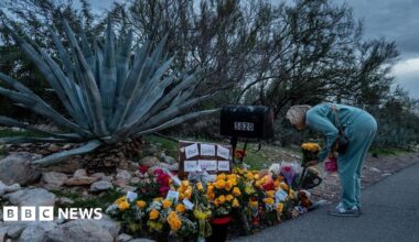 A woman in a teal sweatsuit bends over and lays flowers at a memorial for Nancy Guthrie