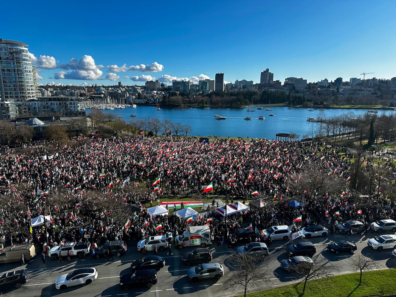 A large crowd gathers at a waterfront park, many holding flags, with the city skyline visible in the background on a clear day.