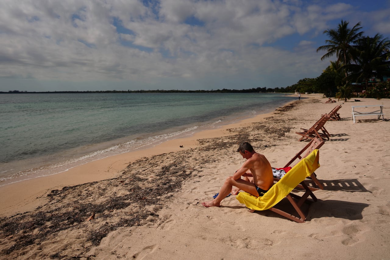 A person sits on a chair covered with a yellow towel on a beach, with the ocean and sand stretching out for a few miles.