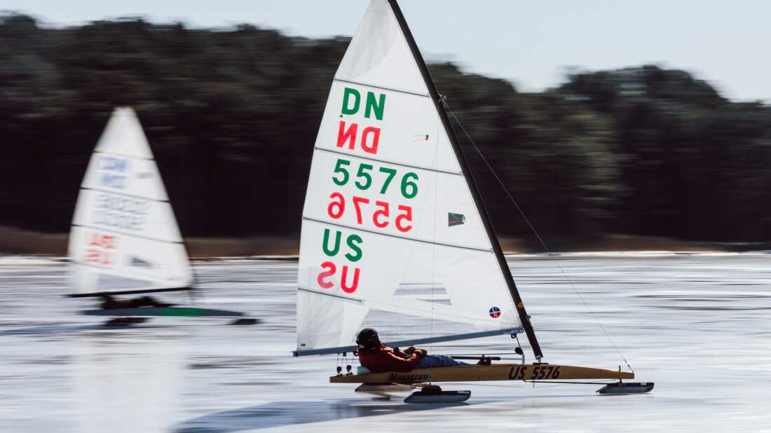Iceboat racer and sailor, Ray Gauthier, sails across the ice in Claiborne, Md. The wooden boats are skinny and have ice skates underneath and a large white sail with colorful numbers and letters. Ray is racing with another ice boat nearby.