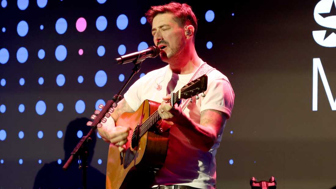 Marcus Mumford, with his eyes closed, sings into a mic and plays a guitar.