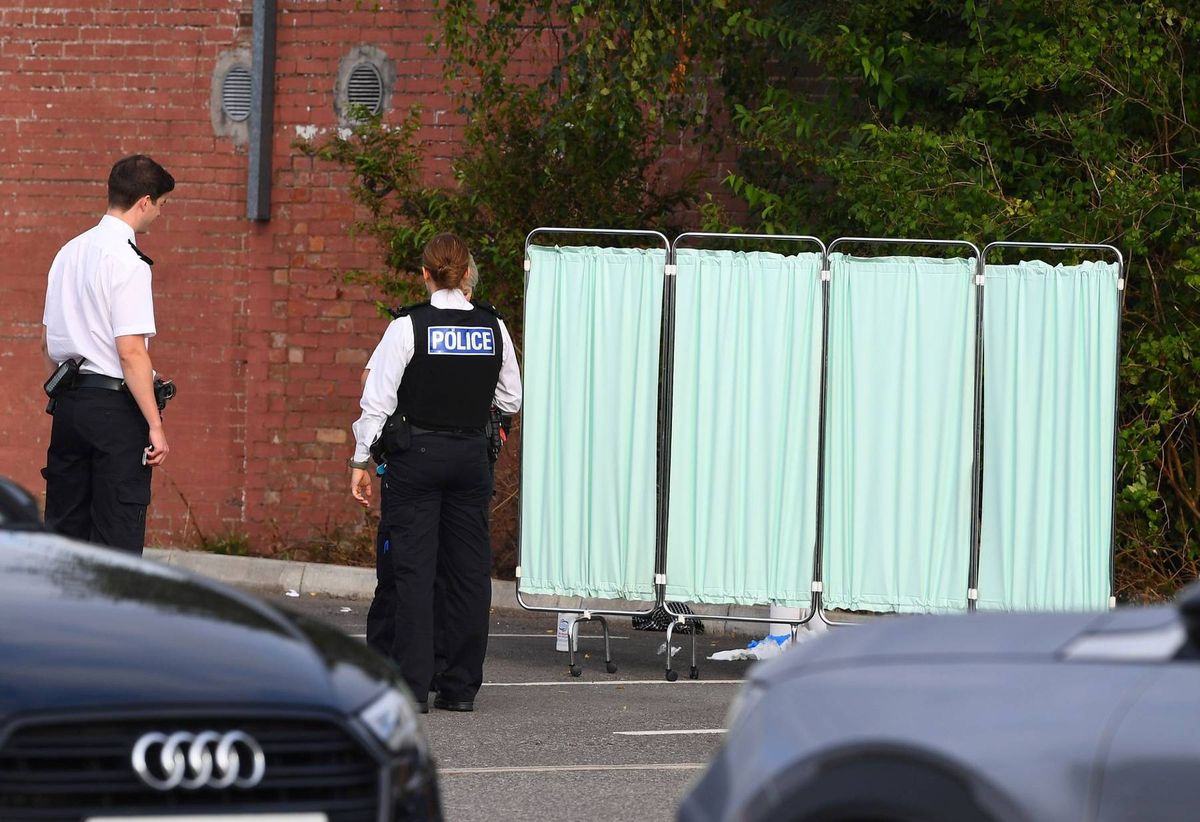 Two individuals dressed in police uniforms are standing near a temporary structure in an outdoor setting. They are positioned next to a black vehicle, with additional cars visible in the background.