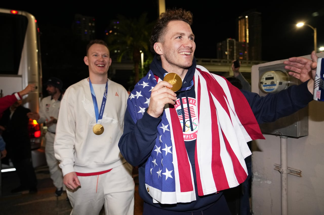 Two men smile while walking in a nighttime scene, with medals around their necks and one with an American flag over their shoulders.