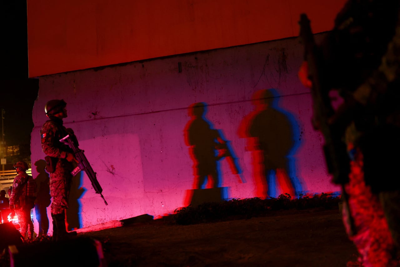 Men with military uniforms and guns stand outside in the glow of a pink light