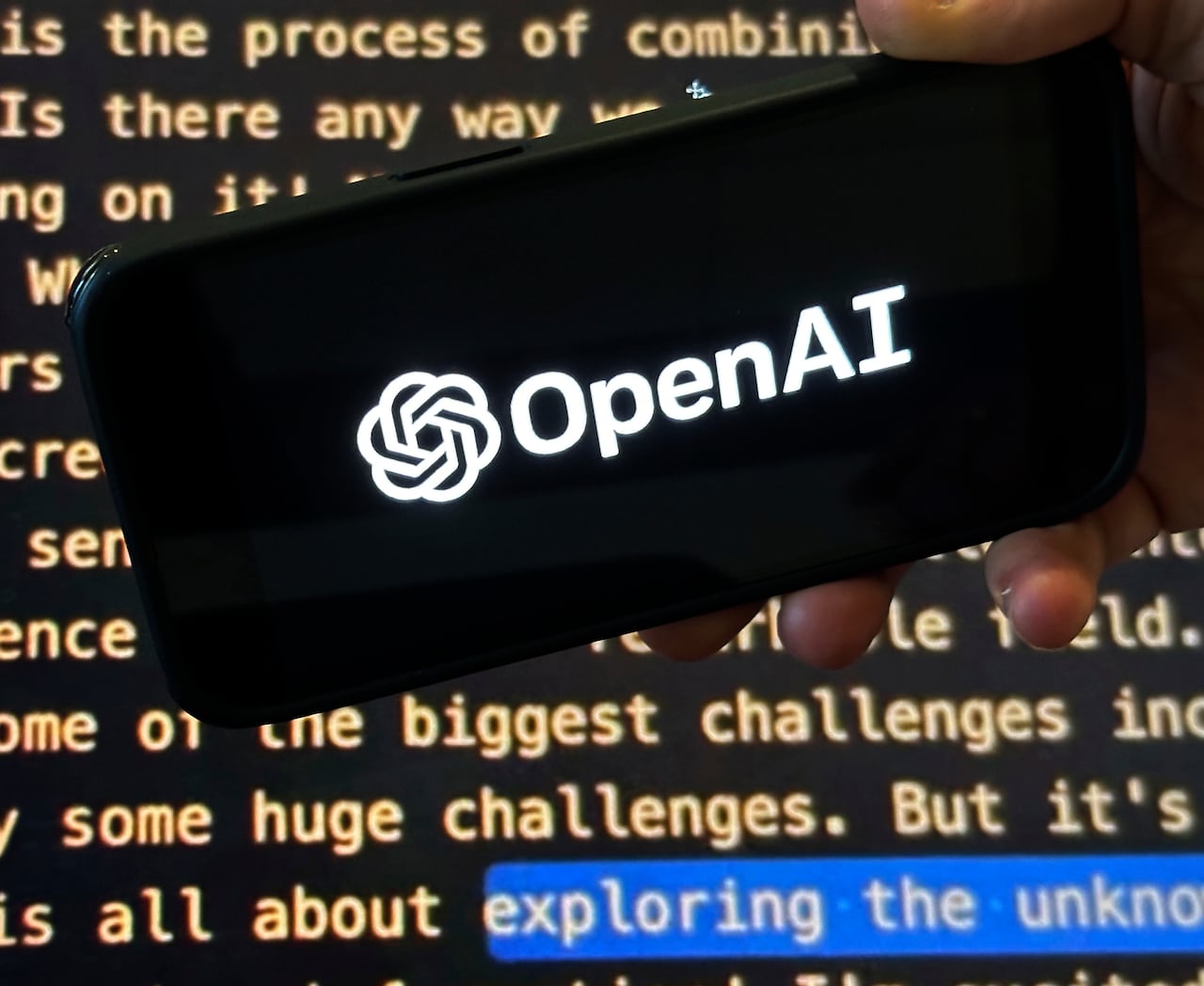 The OpenAI logo appears on a mobile phone in front of a screen showing part of the company website