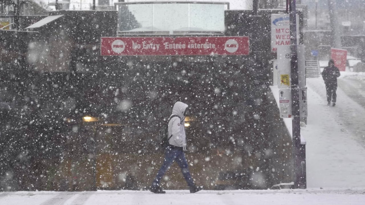 Snow falls on a city street, as a person crosses an intersection.