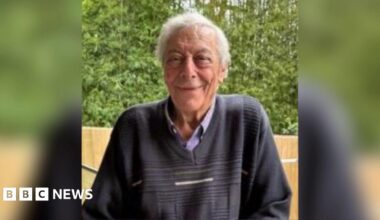 An elderly man with grey hair and wearing a dark blue jumper smiles at the camera