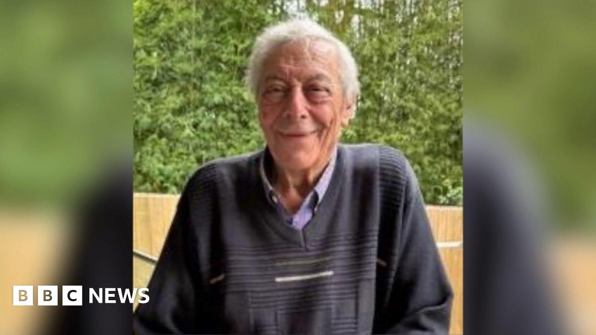 An elderly man with grey hair and wearing a dark blue jumper smiles at the camera