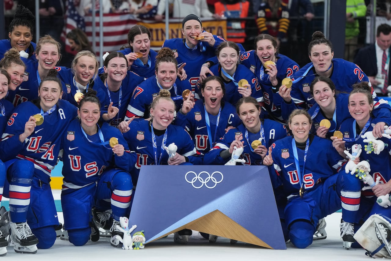 Women in hockey gear smile and hold up gold medals