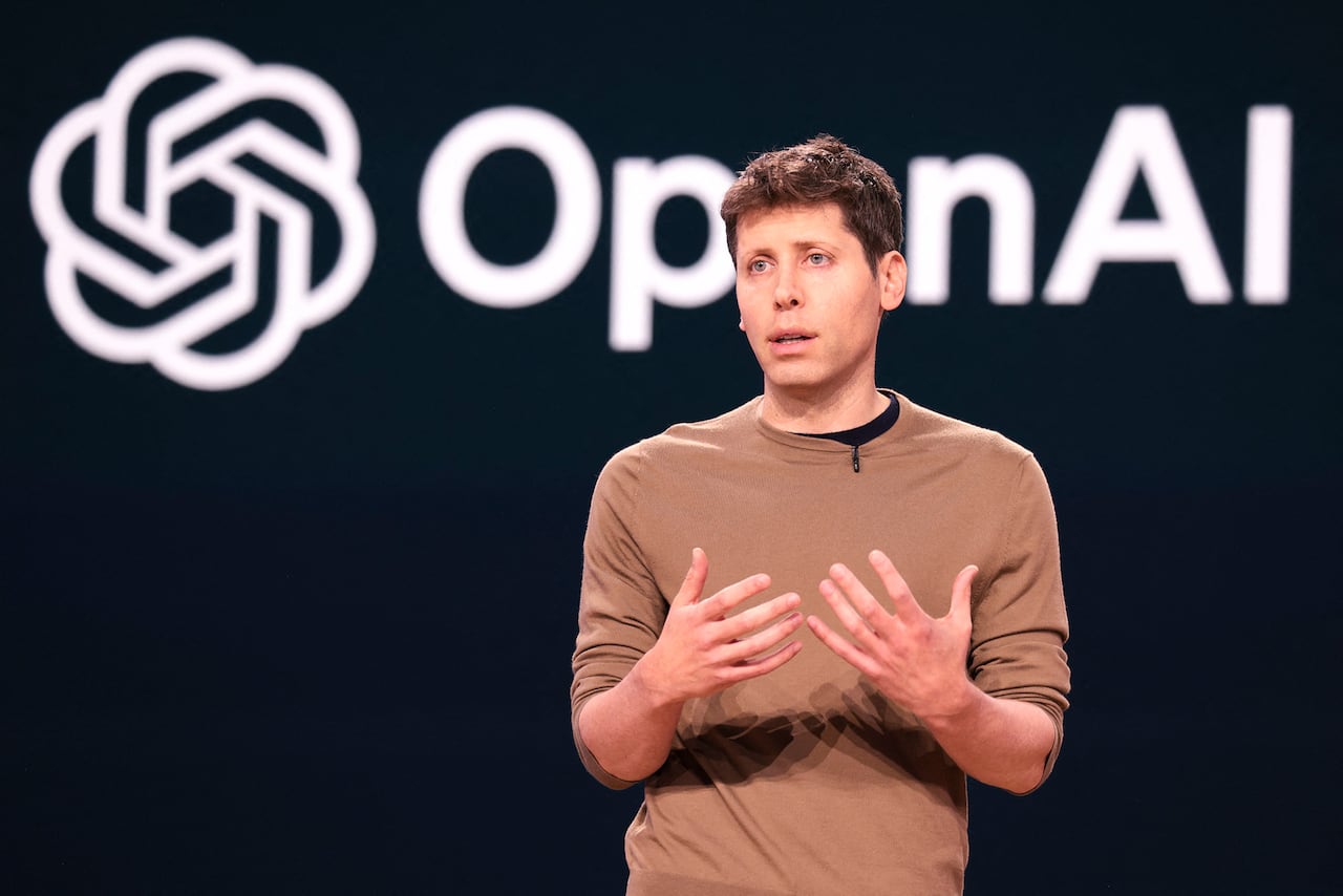 A man stands on a shirt with the words 'OpenAI' on a screen behind him.