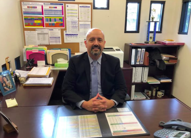 A man with medium-toned skin sits behind a desk with his hands held together. He's wearing a suit and tie and is surrounded by books and papers neatly stacked. 