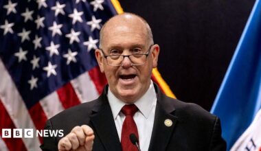 Tom Homan stands outside in front of leafless trees wearing a grey suit.