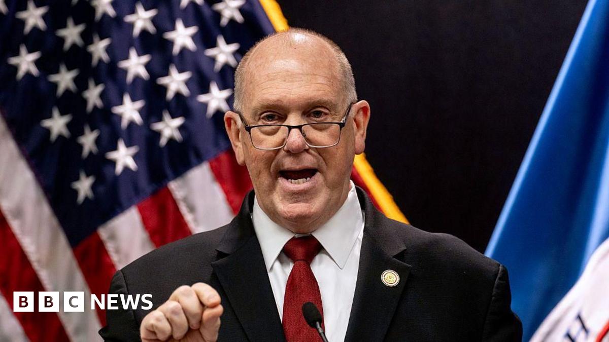 Tom Homan stands outside in front of leafless trees wearing a grey suit.
