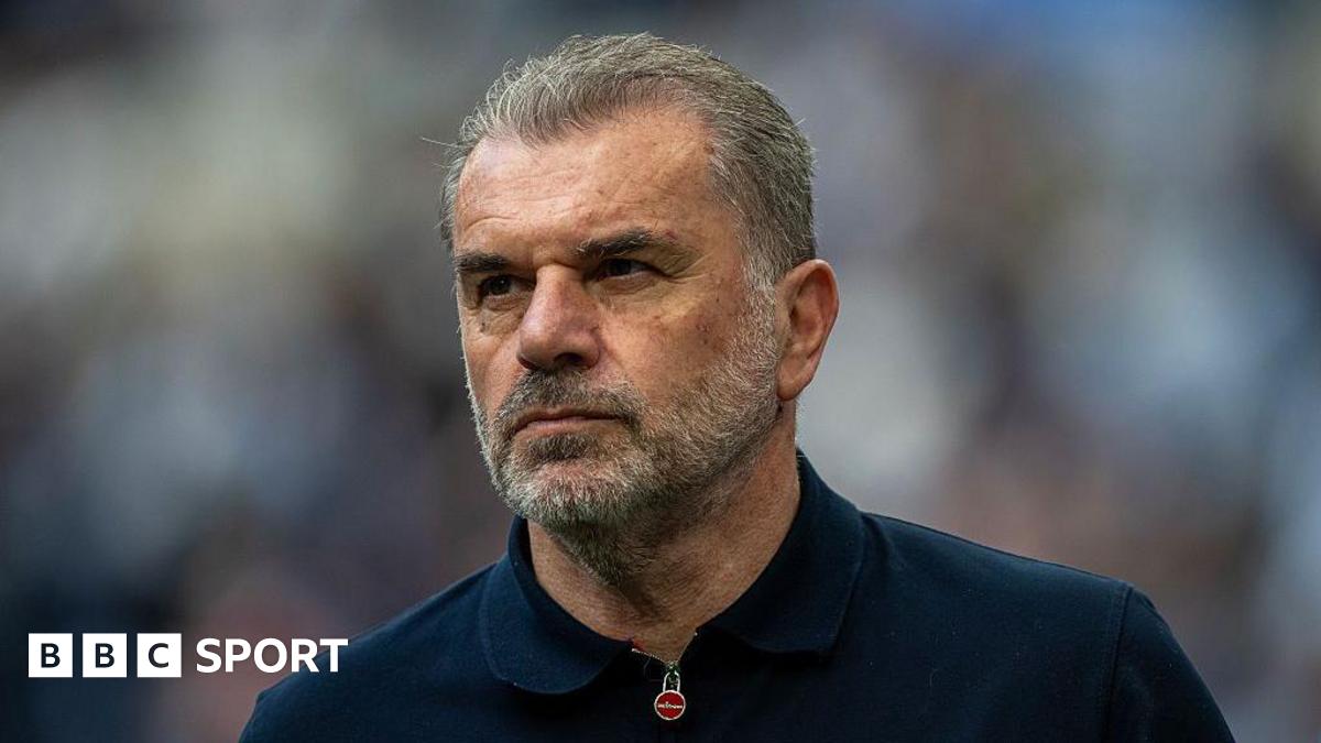 Close-up of Ange Postecoglou. He has short grey hair, brushed away from his face, and a grey beard. He is wearing a navy top which has a zip fastening at the collar.
