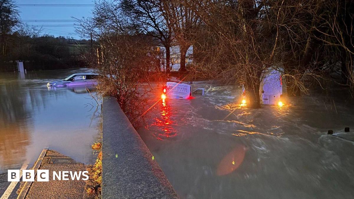 Three vehicles - two vans and a Range Rover - are half submerged in floodwater in Axminster, Devon, during Storm Chandra. The two vans have their lights on. The photo was taken during the early hours of the morning. Several trees and a house are in the background.