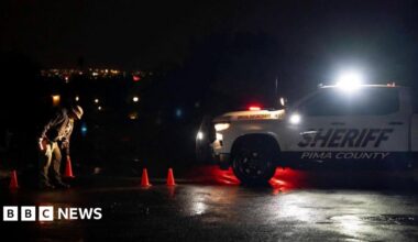 An official examines the ground near orange traffic cones at night, illuminated by headlights from a Pima County Sheriff’s vehicle.