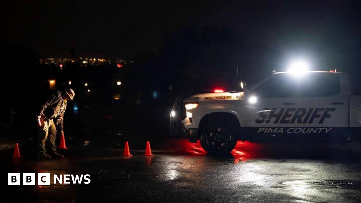 An official examines the ground near orange traffic cones at night, illuminated by headlights from a Pima County Sheriff’s vehicle.