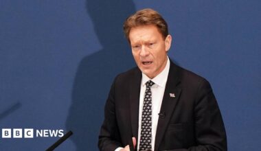 Richard Tice delivering his speech. He's wearing a dark suit, white tie and dark tie with pattern, standing in front of a blue display.