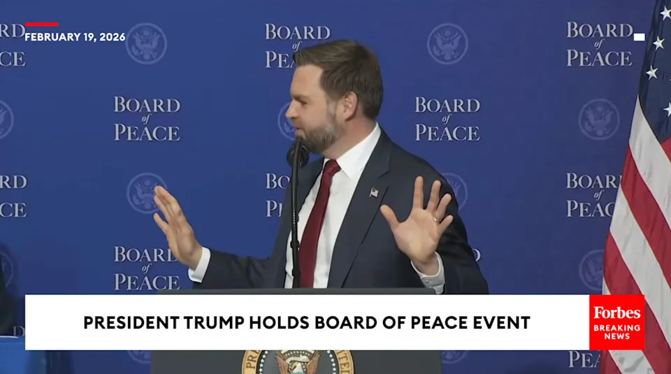 Man in suit and tie gestures during speech at Board of Peace event, February 2026. Event branded backdrop is visible