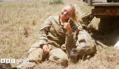A 25-year-old girl with long, blonde hair tied in a ponytail smiles and gives a thumbs up as she sits in a field on a warm summer's day. She is wearing a camouflage Army uniform, and looks to have a light coating of paint or mud on her cheeks. Next to a water bottle, a camouflage backpack, and the rear of a truck-like vehicle.