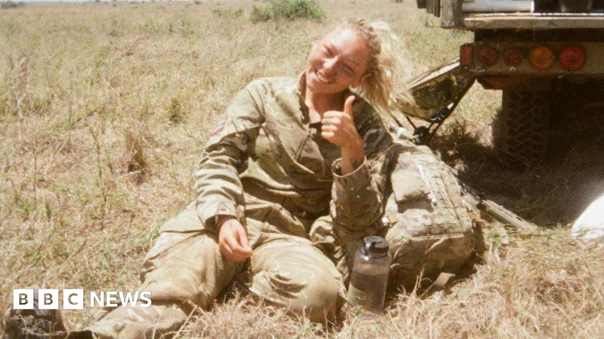A 25-year-old girl with long, blonde hair tied in a ponytail smiles and gives a thumbs up as she sits in a field on a warm summer's day. She is wearing a camouflage Army uniform, and looks to have a light coating of paint or mud on her cheeks. Next to a water bottle, a camouflage backpack, and the rear of a truck-like vehicle.