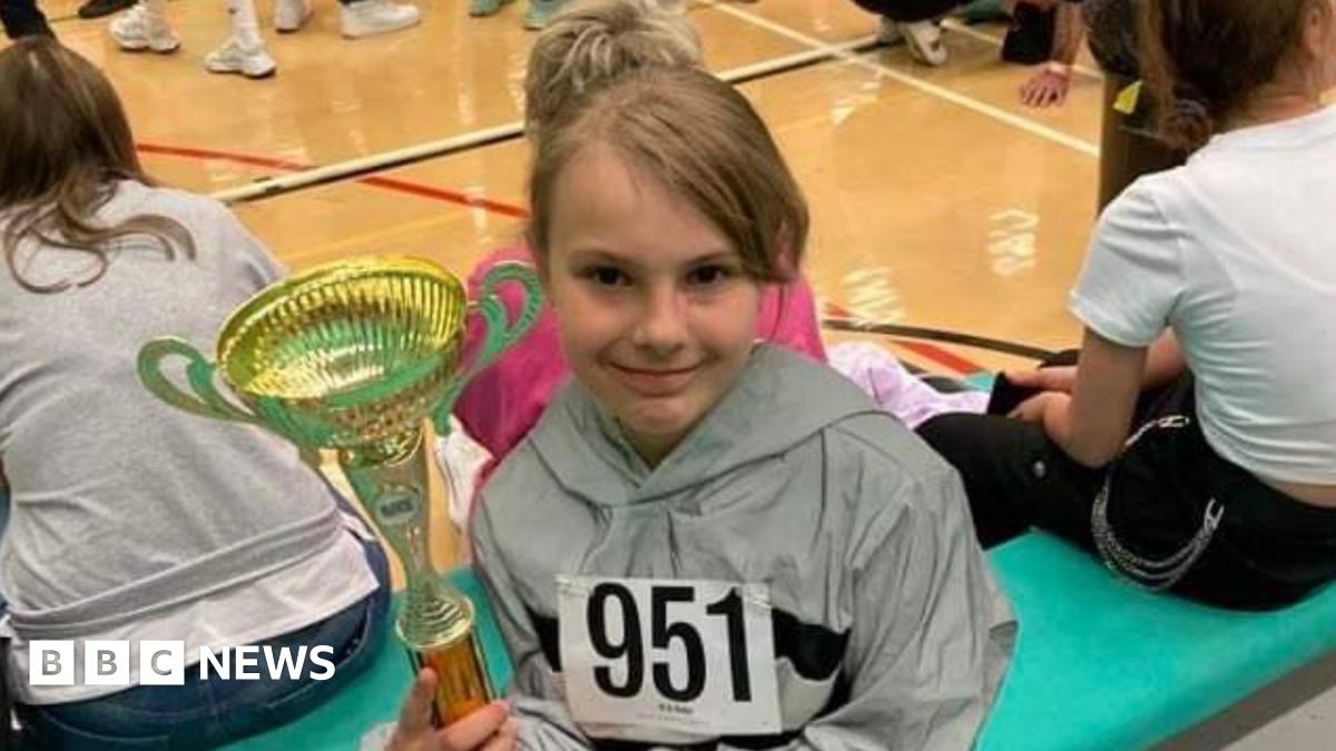 A young girl, her hair tied back, sits on a bench with other girls similar in age to herself. She is holding a trophy.