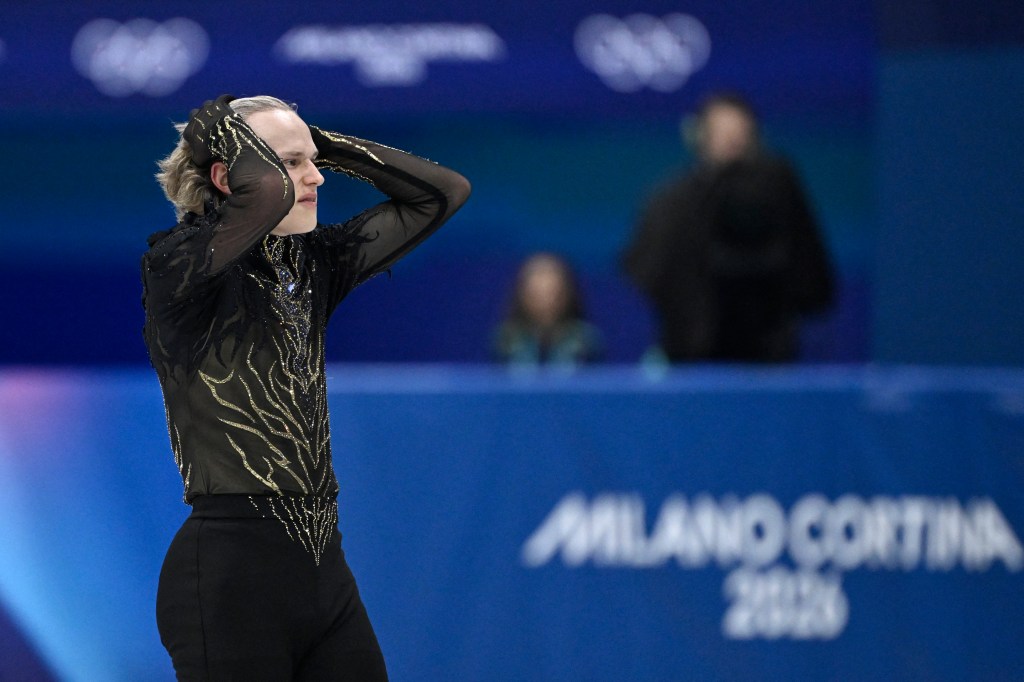 A figure skater with blonde hair in a black costume with gold designs, holding his hands to his head in apparent frustration.