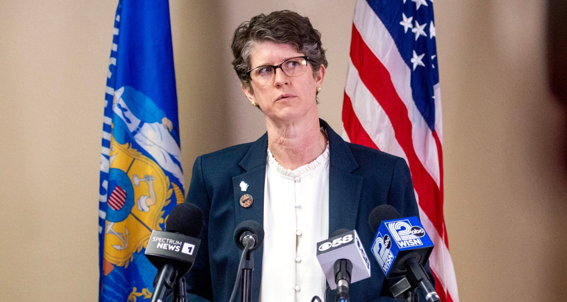 A woman in a suit stands at a podium with microphones, speaking in front of American and Wisconsin state flags.
