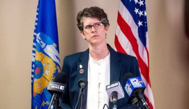 A woman in a suit stands at a podium with microphones, speaking in front of American and Wisconsin state flags.