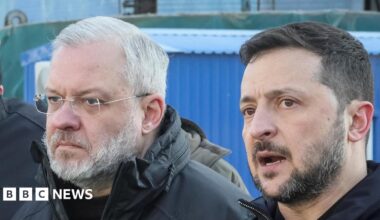 German Galushchenko, with grey hair, beard, and glasses with a neutral expression. He is standing next to Volodymyr Zelensky. Both men are wearing black jackets.