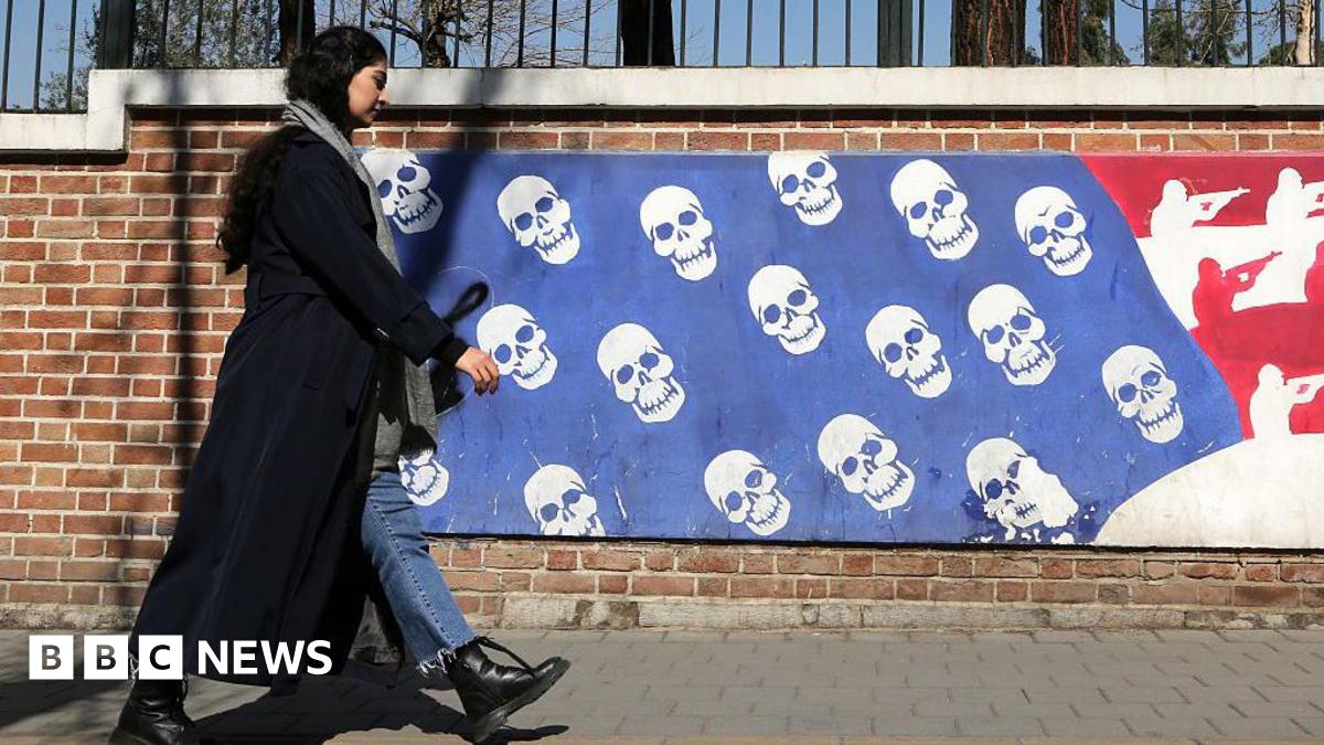 A woman in black in Iran walks near a mural that depicts the US flag with skulls instead of stars