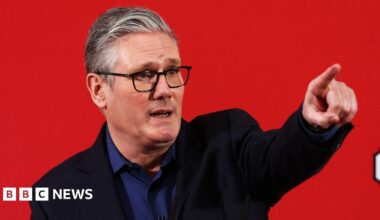 Keir Starmer in a blue shirt and black blazer standing in front of a red background pointing into the distance.