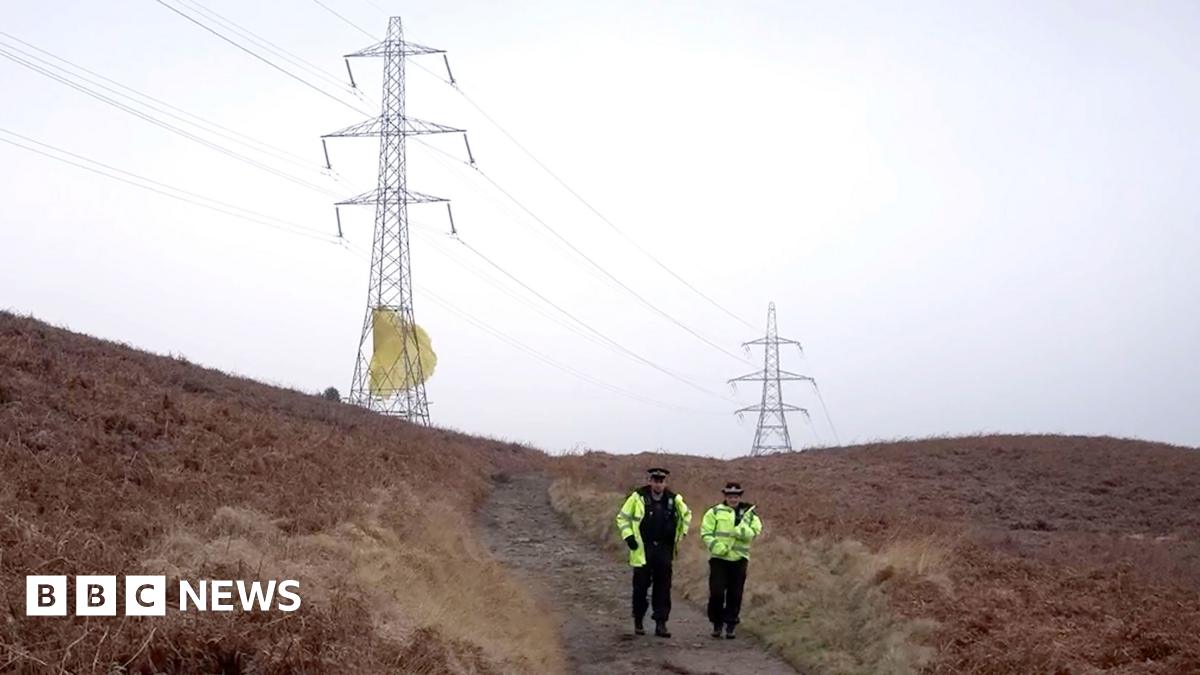 A male and a female police officer walk down a remote dirt track, which is flanked on both side by gorse. On the horizon are two large electricity pylons, there is yellow-coloured material that is wrapped around the bottom of the nearest pylon that appears to be billowing in the wind