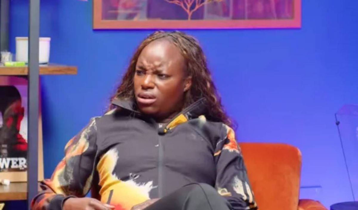 An individual with curly hair is seated on an orange chair against a blue backdrop, appearing to be engaged in a conversation.