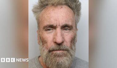Police custody image of Tony Devenport with dishevelled grey hair and beard wearing a grey top.