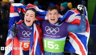 Great Britain's Matt Weston and Tabitha Stoecker, both wearing Team GB lycra kit and draped in a union flag, punch the air as they celebrate winning gold after the Skeleton Mixed Team Final at the Cortina Sliding Centre, on day nine of the Milano Cortina 2026 Winter Olympics, Italy.