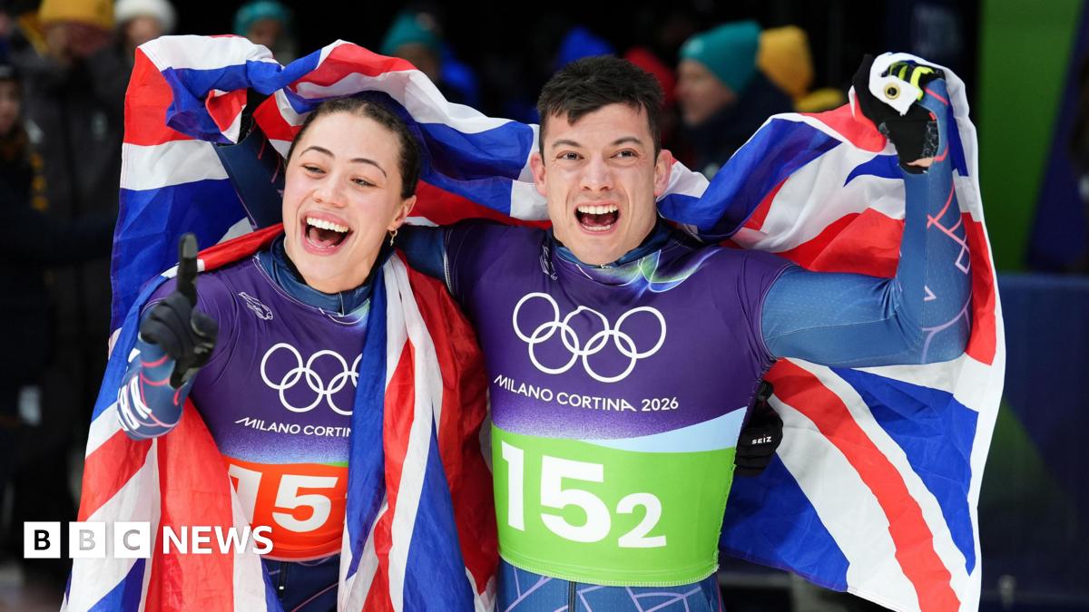 Great Britain's Matt Weston and Tabitha Stoecker, both wearing Team GB lycra kit and draped in a union flag, punch the air as they celebrate winning gold after the Skeleton Mixed Team Final at the Cortina Sliding Centre, on day nine of the Milano Cortina 2026 Winter Olympics, Italy.