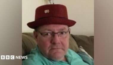 A grainy photograph of Gary Piatek, a white, middle-aged man wearing a red hat and a green polo shirt. He is sitting on a sofa.