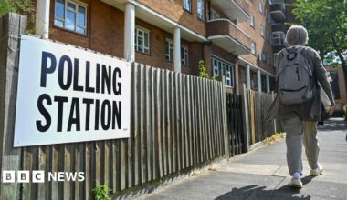 A white sign with polling station written in black text is tied to a wood panelled fence in front of a block of flats. A person wearing a backpack is walking down the pavement.