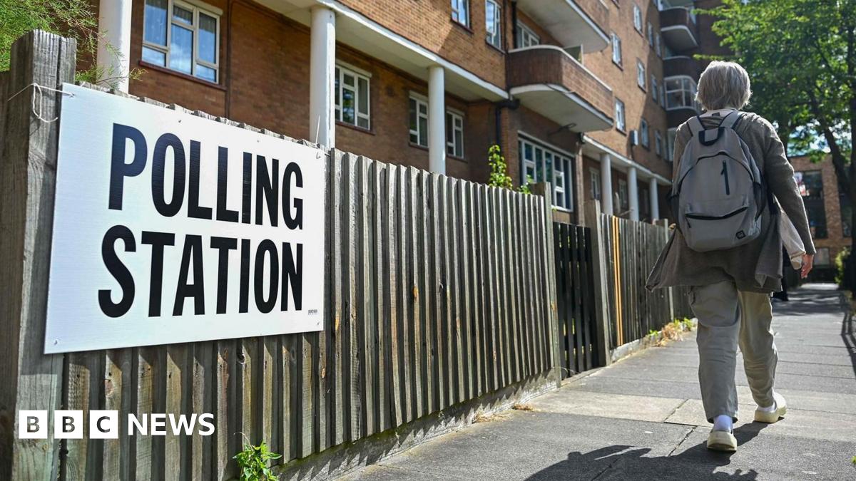 A white sign with polling station written in black text is tied to a wood panelled fence in front of a block of flats. A person wearing a backpack is walking down the pavement.