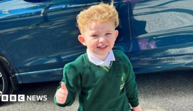 Peter Maughan, a four-year-old boy with short, blond hair, is standing by a blue car wearing a school uniform. He has a white shirt, a green and yellow tie and a green jumper. He is holding a green schoolbag and is making a thumbs-up sign.