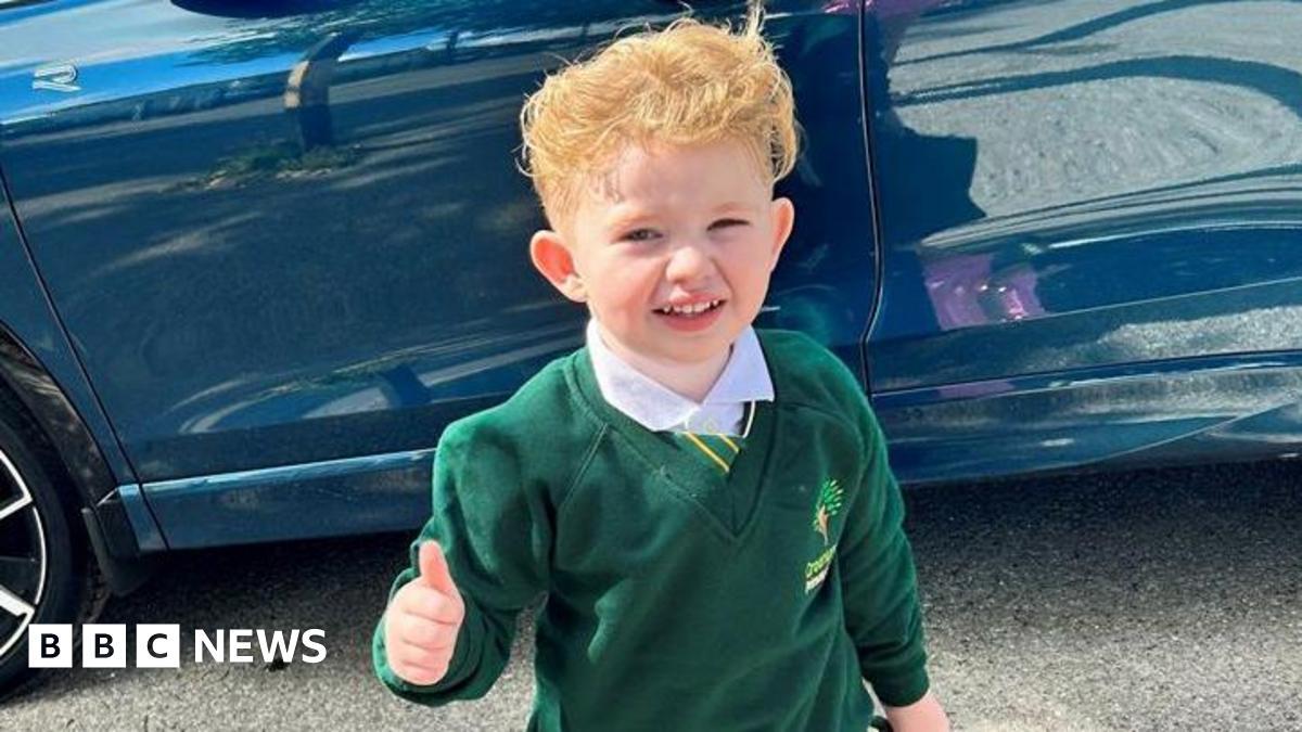 Peter Maughan, a four-year-old boy with short, blond hair, is standing by a blue car wearing a school uniform. He has a white shirt, a green and yellow tie and a green jumper. He is holding a green schoolbag and is making a thumbs-up sign.