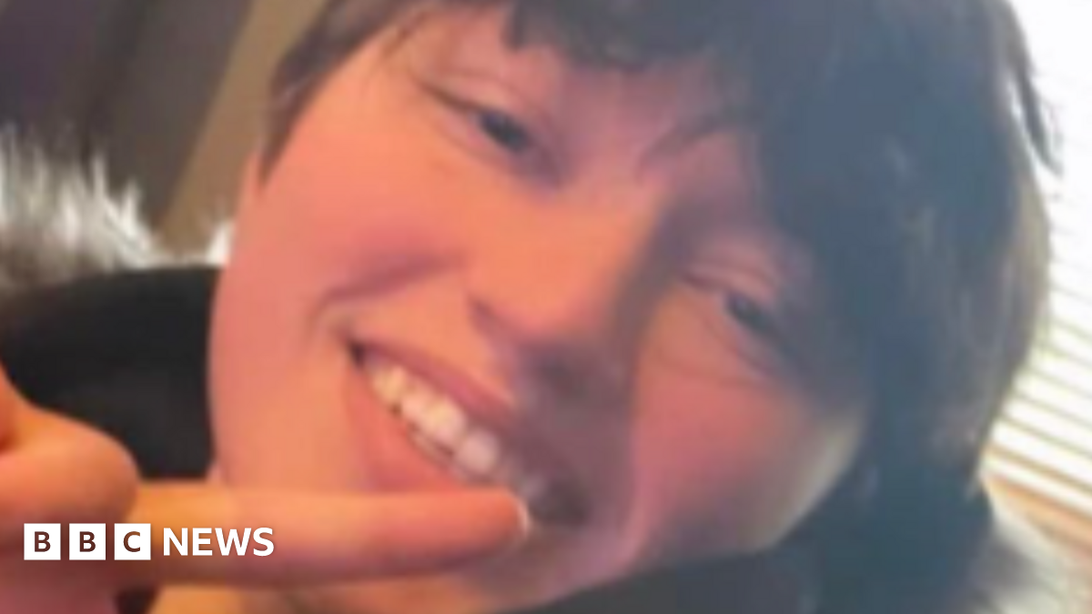 Close up picture of Demi smiling looking at the camera. She has short brown hair and wears a black coat with a fur hood. She is doing a peace pose with her one hand.