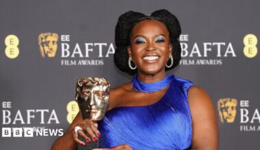 Wunmi Mosaku grins as she holds her Bafta award in front of the films awards backdrop. She wears a blue off shoulder dress and scarf.