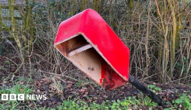 A view of a damage postbox. The pole that the box is on is at an angle and the front door of the postbox is missing.