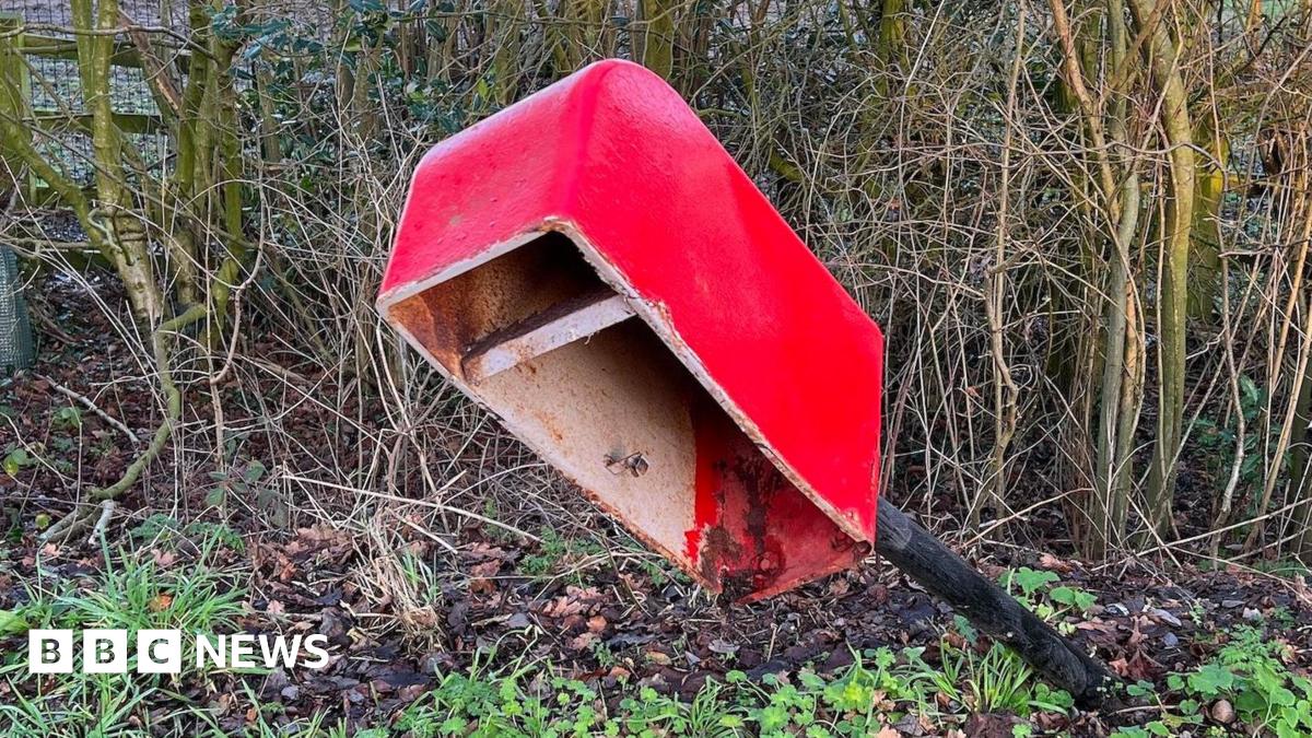 A view of a damage postbox. The pole that the box is on is at an angle and the front door of the postbox is missing.